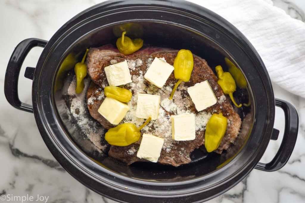 overhead of a Mississippi pot roast in a slow cooker before it is cooked, pads of butter and seasoning visible on top