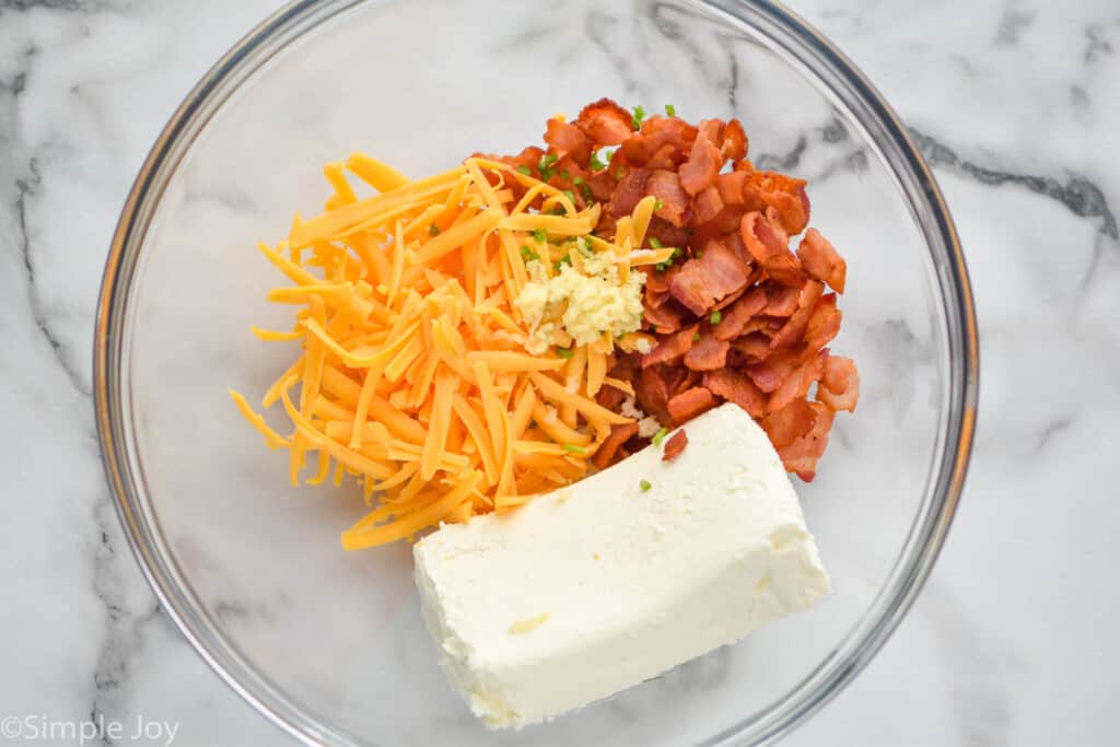 overhead of a bowl of cream cheese, shredded cheddar cheese, seasonings, and cooked bacon pieces