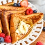 four triangles of French toast recipe sitting on a white plate with butter, syrup, and fresh berries