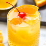 close up of an amaretto stone sour recipe in a glass tumbler with ice, an orange slice, and a cherry