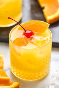 close up of an amaretto stone sour recipe in a glass tumbler with ice, an orange slice, and a cherry