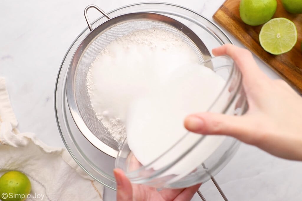 a metal sieve propped on a glass bowl with flour in it, and sugar being poured into it from a bowl