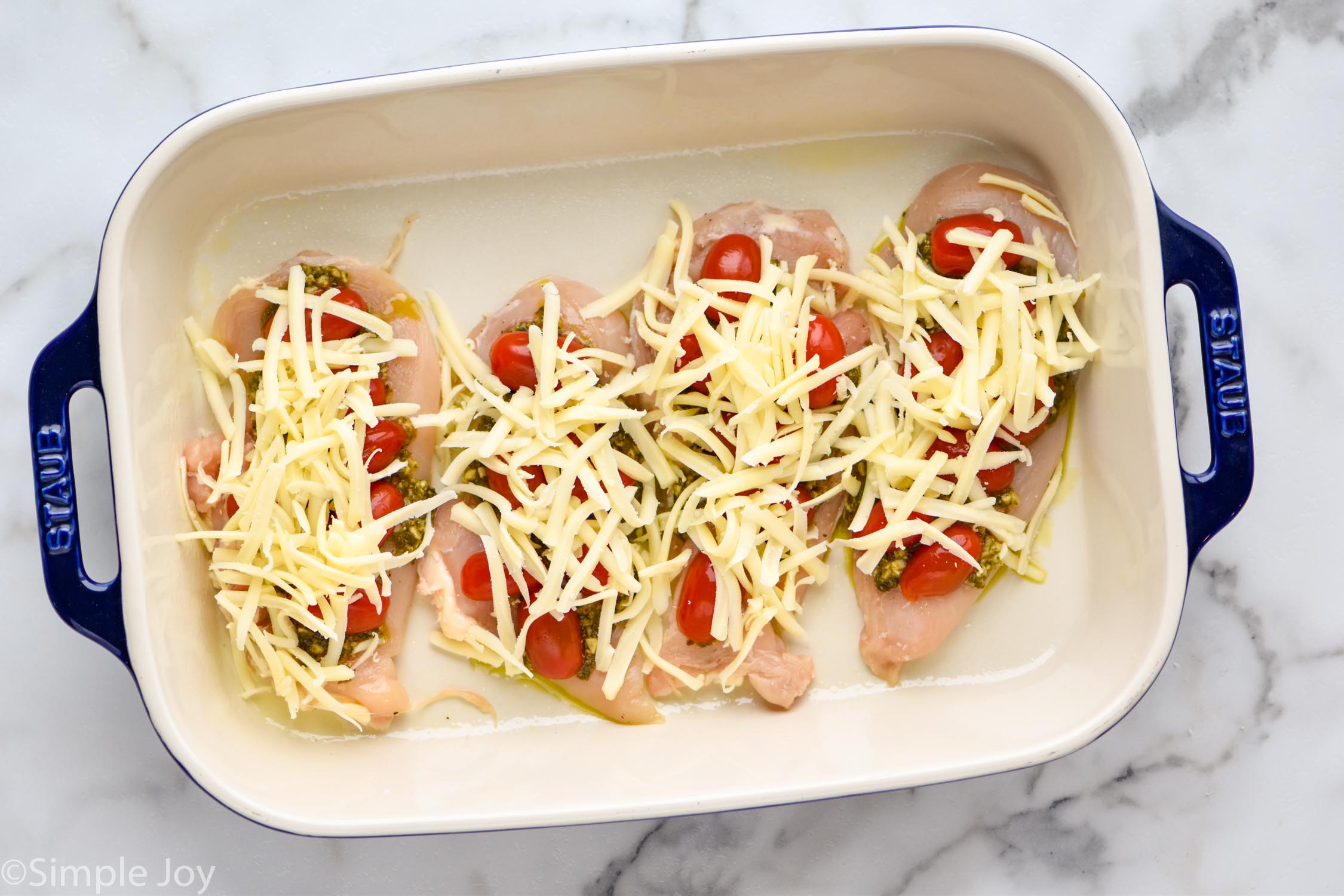overhead of a baking dish will four chicken cutlets with pesto, cherry tomatoes, and shredded mozzarella cheese