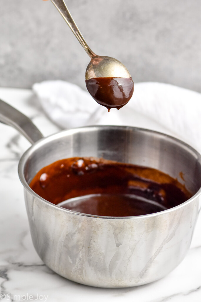 small sauce pan of hot fudge recipe with a spoon above it, sauce coating the spoon