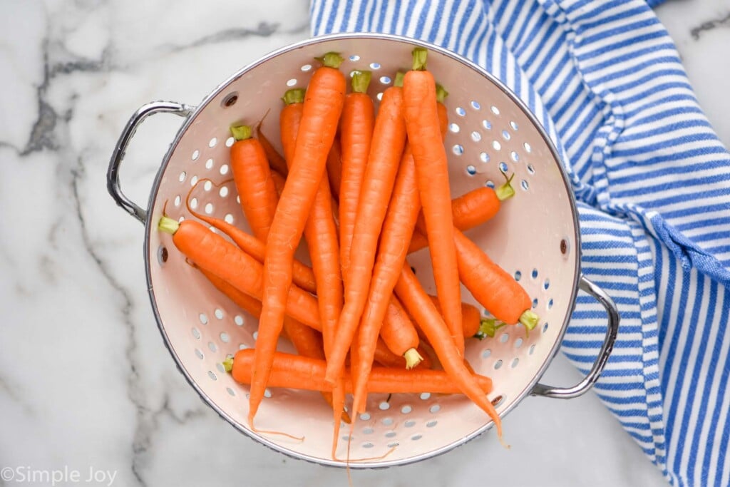 Whole carrots in white strainer next to a blue and white striped towel.