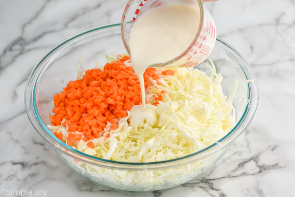 pouring coleslaw dressing over a bowl of cut cabbage and diced carrots