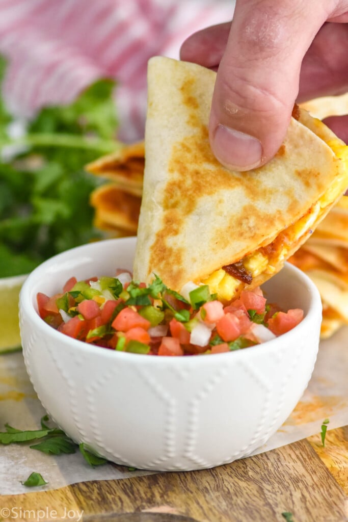 Photo of man's hand dipping a slice of breakfast quesadilla into pico de gallo as garnish.