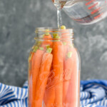 Clear mason jar filled with whole carrots. Measuring cup above jar pouring clear liquid into jar to make pickled carrots. - 10