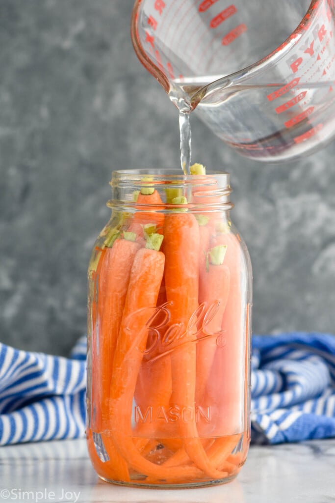 Clear mason jar filled with whole carrots. Measuring cup above jar pouring clear liquid into jar to make pickled carrots.