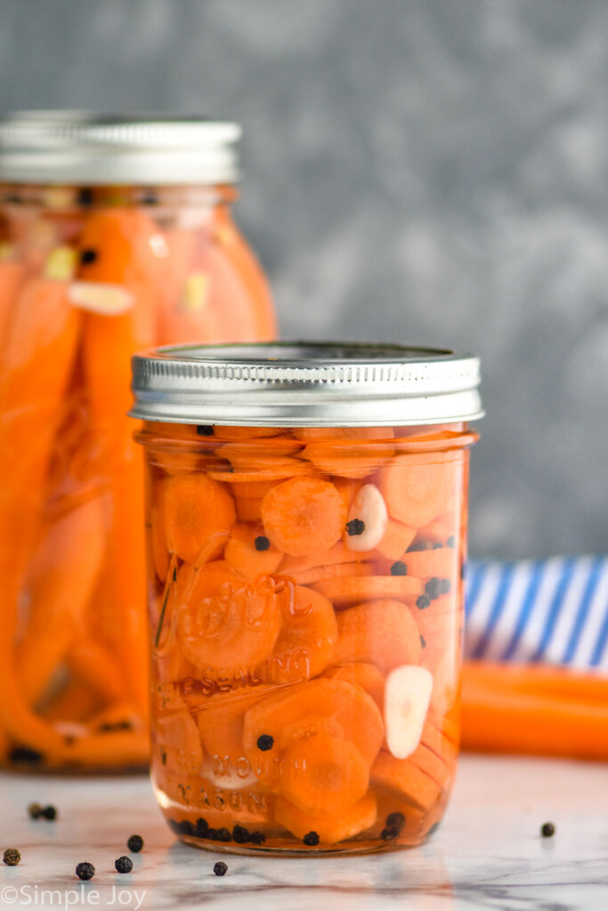 Photo of pickled carrots recipe. Sealed mason jar filled with diced pickled carrots sitting on counter top with large jar of whole pickled carrots behind it.