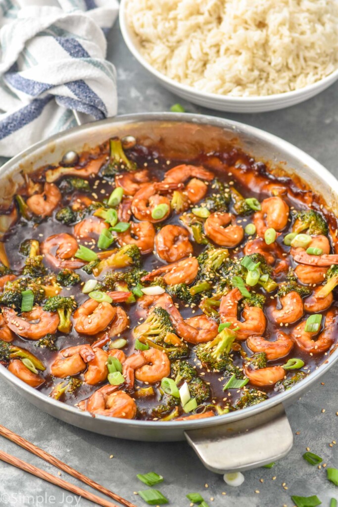 Overhead photo of shrimp and broccoli stir fry in a stainless steel pan. Bowl of white rice beside the pan.