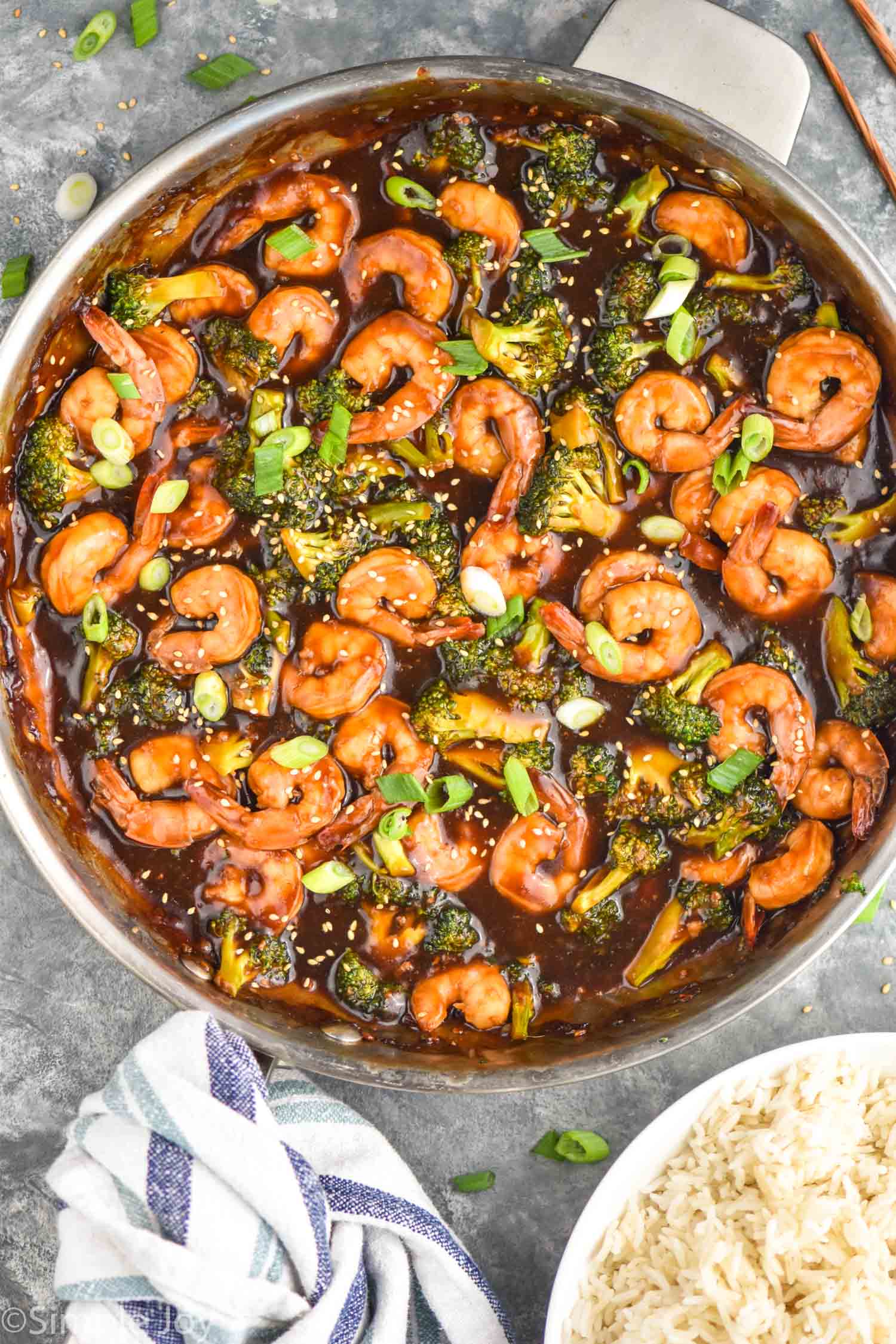 Overhead photo of shrimp and broccoli stir fry in a stainless steel pan. Bowl of white rice beside the pan.