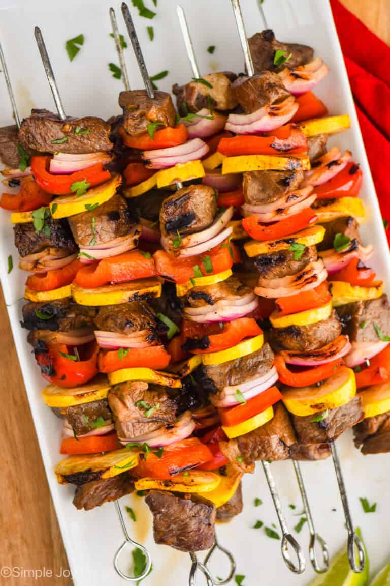 a pile of steak kabobs made with red peppers, yellow zucchini, and red onions with metal skewers on a white plate and garnished with parsley