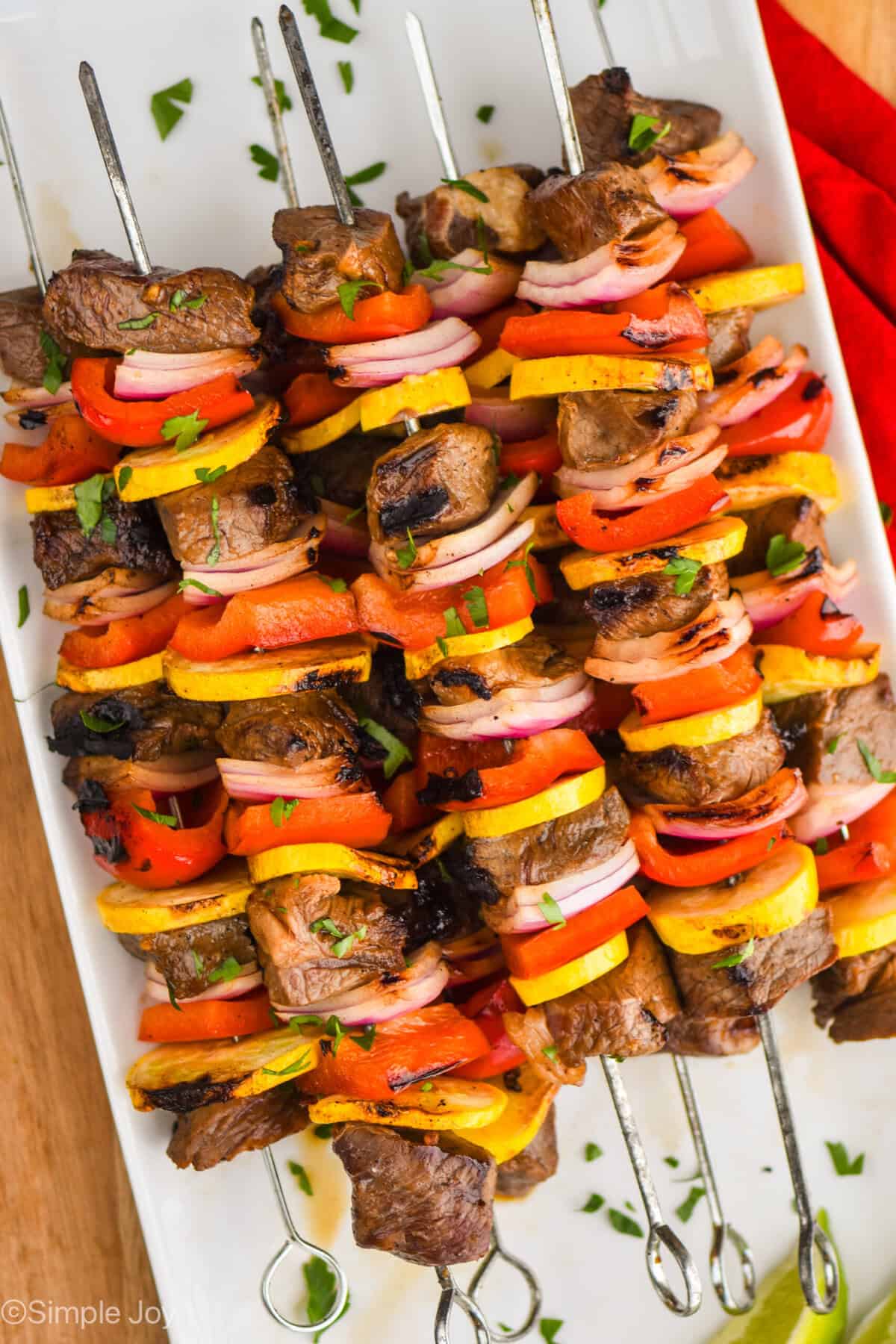 a pile of steak kabobs made with red peppers, yellow zucchini, and red onions with metal skewers on a white plate and garnished with parsley