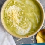 Pinterest graphic of zucchini soup recipe. Overhead photo of Zucchini Soup garnished with parmesan cheese and cream. Slice of bread and a spoon on counter beside soup bowl. Text says, "Amazing Zucchini Soup simplejoy.com"