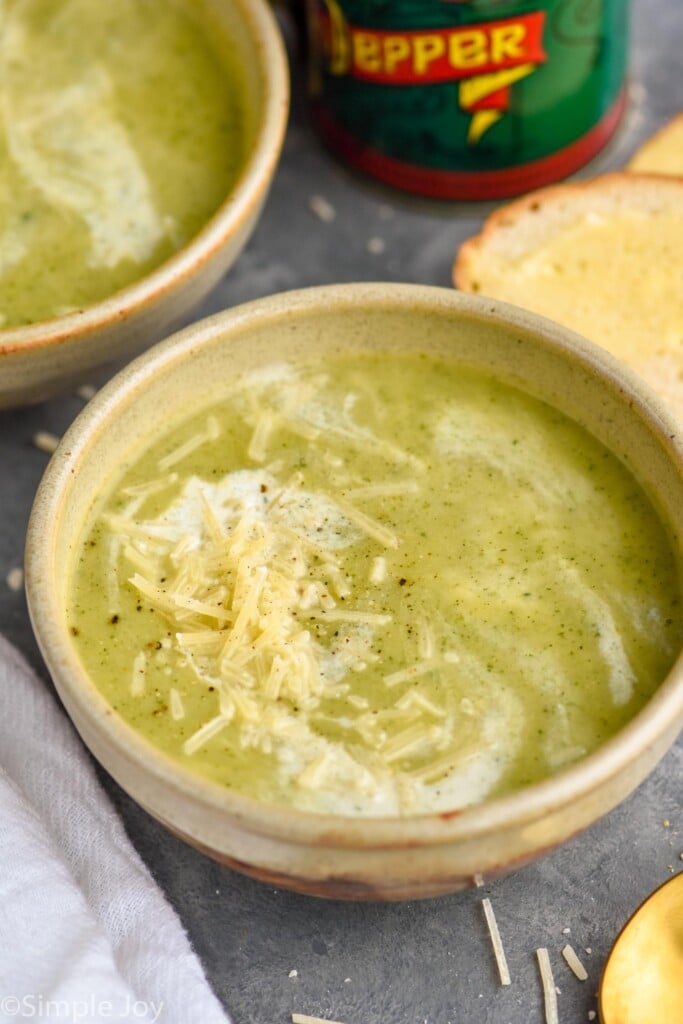 Overhead photo of Zucchini Soup garnished with parmesan cheese and cream. Slice of bread and a spoon on counter beside soup bowl.