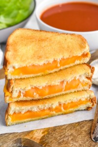Photo of stack of air fryer grilled cheese sandwiches halves with a bowl of tomato soup behind them.