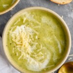 Overhead photo of Zucchini Soup garnished with parmesan cheese and cream. Slice of bread and a spoon on counter beside soup bowl. - 8