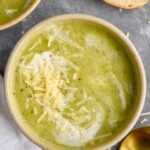 Overhead photo of Zucchini Soup garnished with parmesan cheese and cream. Slice of bread and a spoon on counter beside soup bowl.