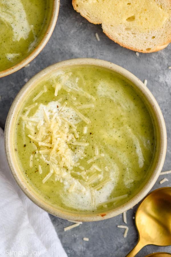Overhead photo of Zucchini Soup garnished with parmesan cheese and cream. Slice of bread and a spoon on counter beside soup bowl.