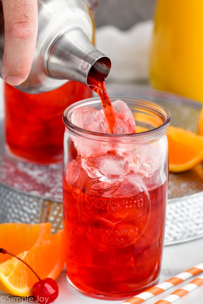 Photo of man's hand pouring Alabama Slammer cocktail into a glass of ice. Pitcher of orange juice in the background and orange slices and cherries sit beside the glass for garnish.