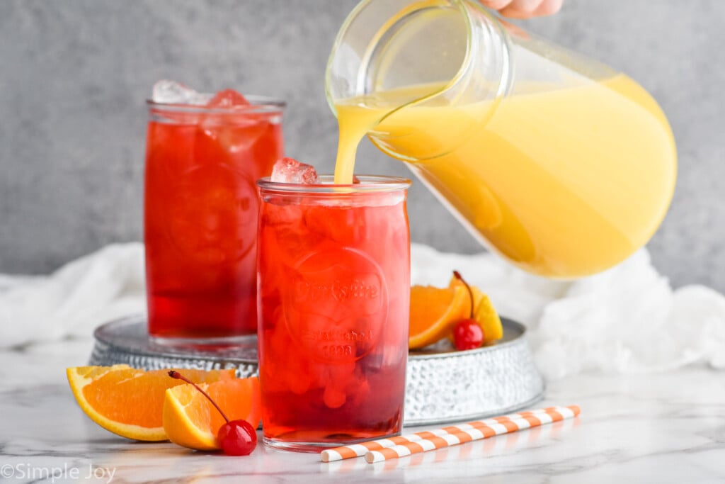 Photo of Alabama Slammer cocktail in two glasses. Pitcher of orange juice is being poured into the first glass. Orange slices, cherries, and straws are on the counter beside the glasses.