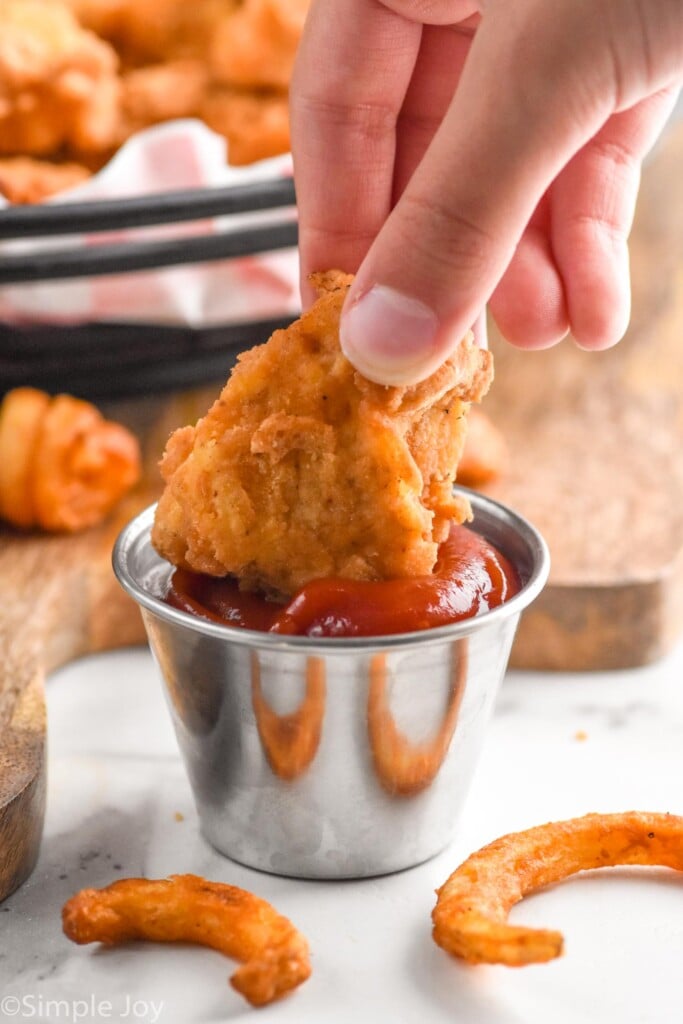 Photo of person's hand dipping a chicken nugget into ketchup. Curly fries on the counter beside the ketchup.