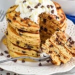 Pinterest graphic for chocolate chip pancakes recipe. Overhead photo of a stack of Chocolate Chip Pancakes garnished with whipped cream and extra chocolate chips. Fork with a large bite of pancakes that has been cut out of stack of pancakes rests on plate. A cup of coffee and bowl of strawberries sit in the background. Text says, "The best chocolate chip pancakes simplejoy.com."