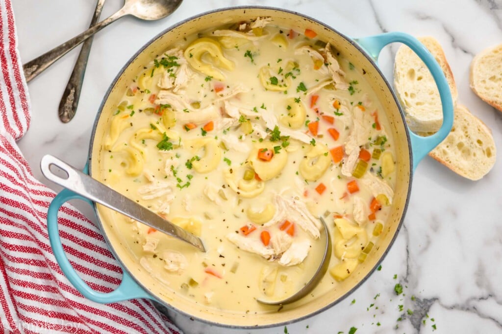 overhead view of pot of chicken tortellini soup with ladle for serving. Spoons and pieces of bread laying beside