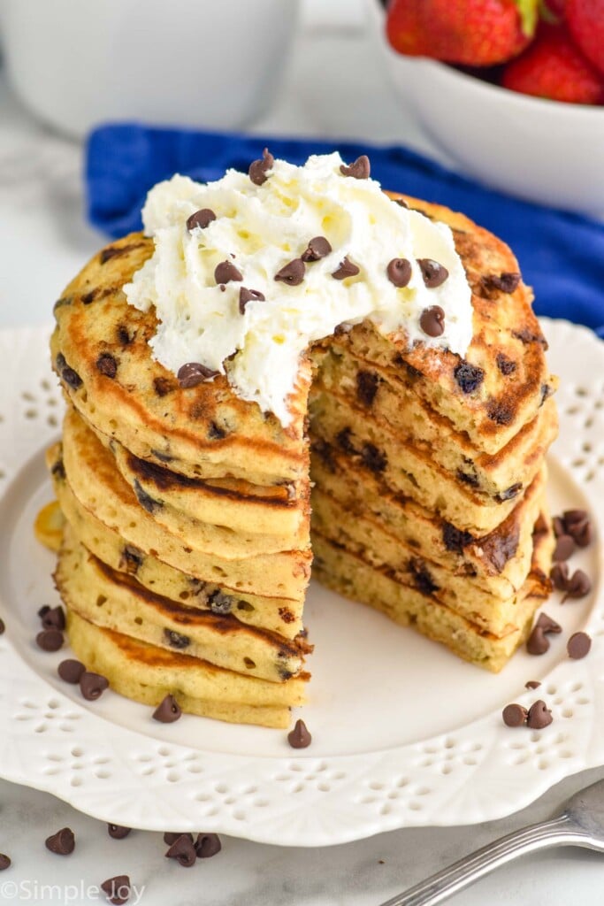 Overhead photo of a stack of Chocolate Chip Pancakes garnished with whipped cream and extra chocolate chips. Large bite has been cut out of stack of pancakes.