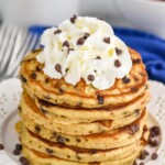 Overhead photo of a stack of Chocolate Chip Pancakes garnished with whipped cream and extra chocolate chips. A cup of coffee and bowl of strawberries sit in the background.