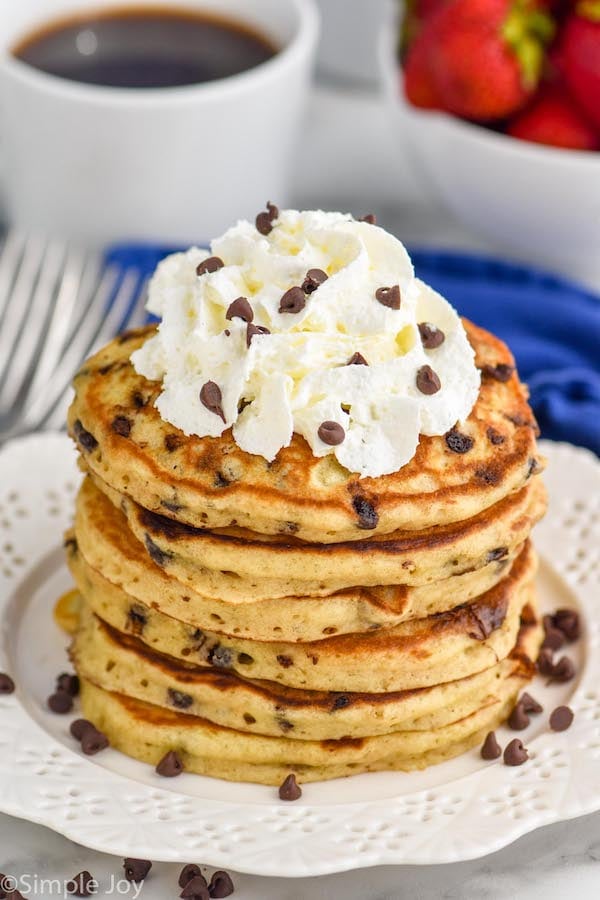 Overhead photo of a stack of Chocolate Chip Pancakes garnished with whipped cream and extra chocolate chips. A cup of coffee and bowl of strawberries sit in the background.
