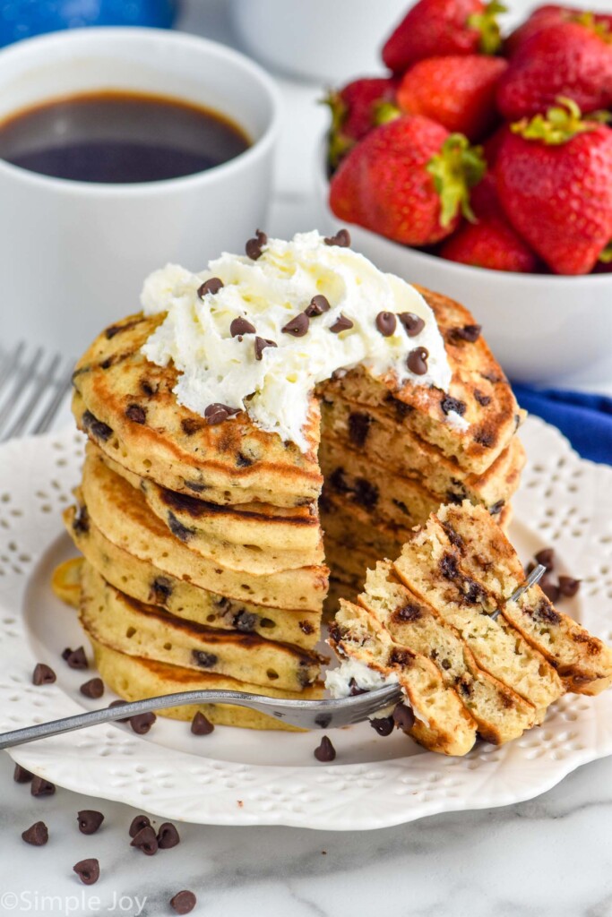 Overhead photo of a stack of Chocolate Chip Pancakes garnished with whipped cream and extra chocolate chips. Fork with a large bite of pancakes that has been cut out of stack of pancakes rests on plate. A cup of coffee and bowl of strawberries sit in the background.