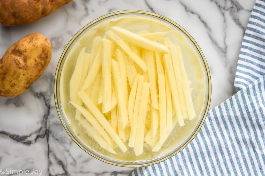 Overhead photo of potato slices soaking in hot water in preparation for easy french fries recipe.