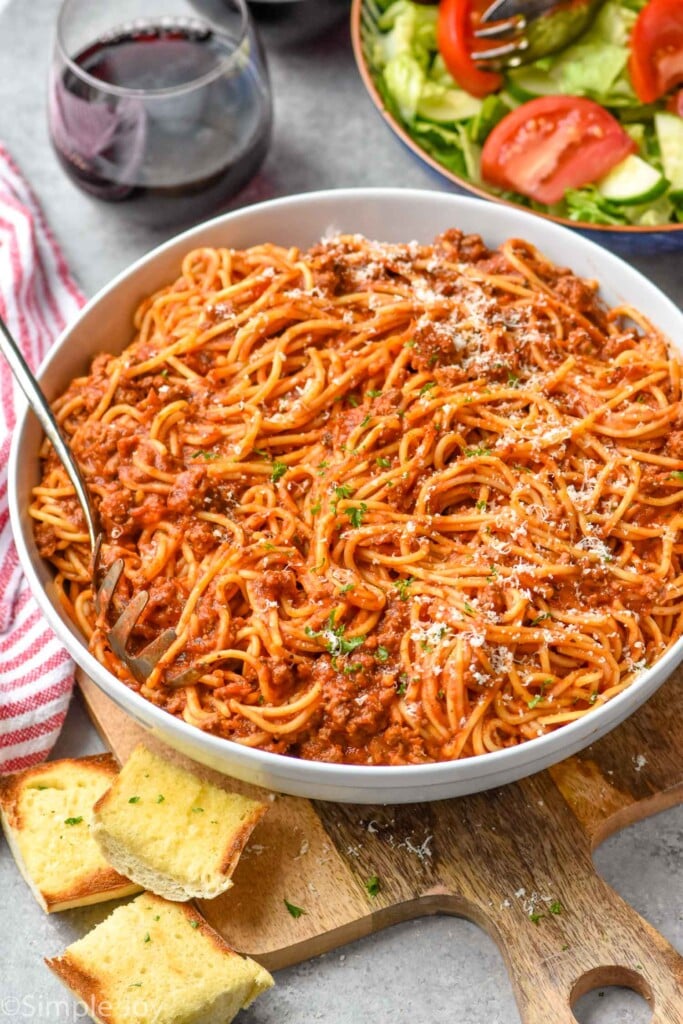 Overhead photo of Crock Pot Spaghetti recipe served in a bowl with parmesan cheese and a fork. Side salad, a glass of wine, and bread sit on counter beside serving bowl.