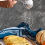 Pinterest graphic for pumpkin cheesecake braid. Text says "super easy pumpkin braid simplejoy.com" Image shows man's hand shaking powdered sugar over a prepared pumpkin cheesecake braid