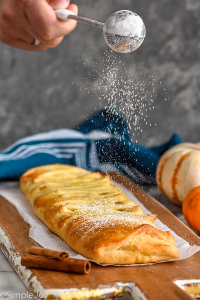 man's hand shaking powered sugar over prepared pumpkin cheesecake braid