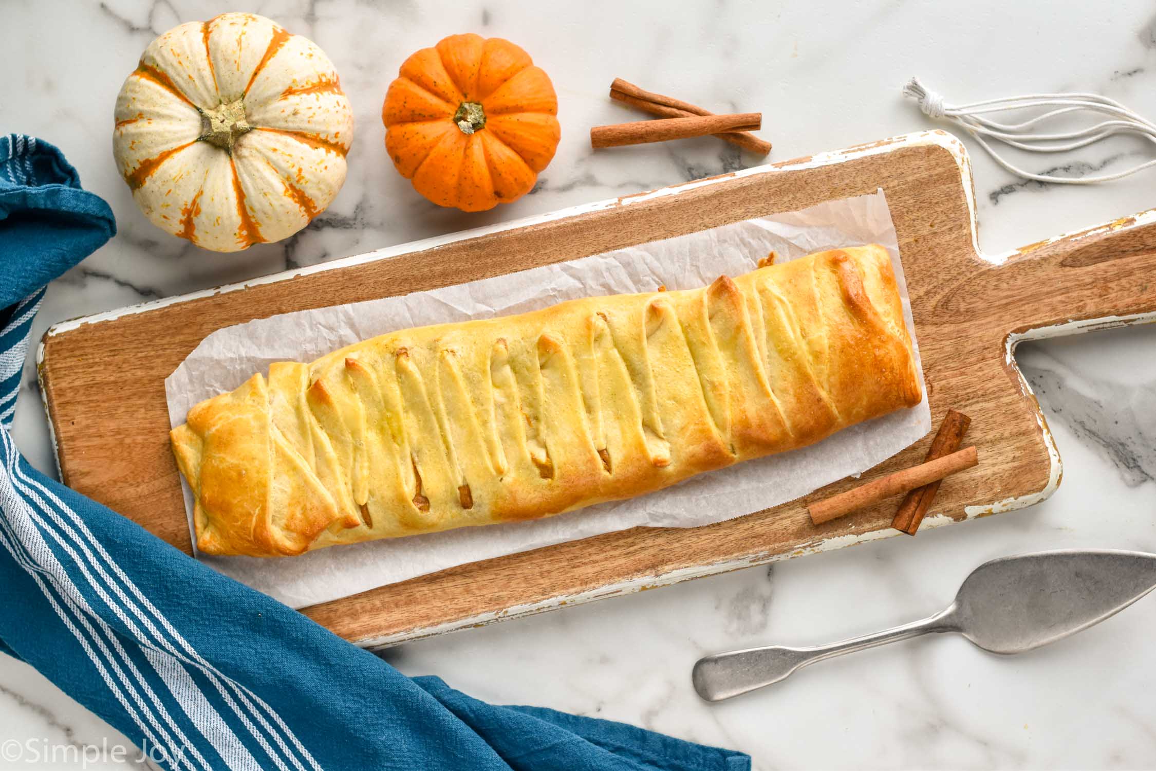 Overhead photo of Pumpkin Cheesecake Braid on a serving board. Pumpkins, cinnamon sticks, and a serving utensil lay on counter beside serving board.