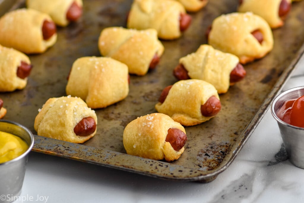baking sheet of pigs in a blanket with containers of ketchup and mustard sitting beside