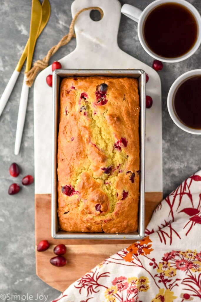 overhead of orange cranberry bread in bread pan sitting on cutting board for serving. Two mugs of coffee and cranberries sitting beside
