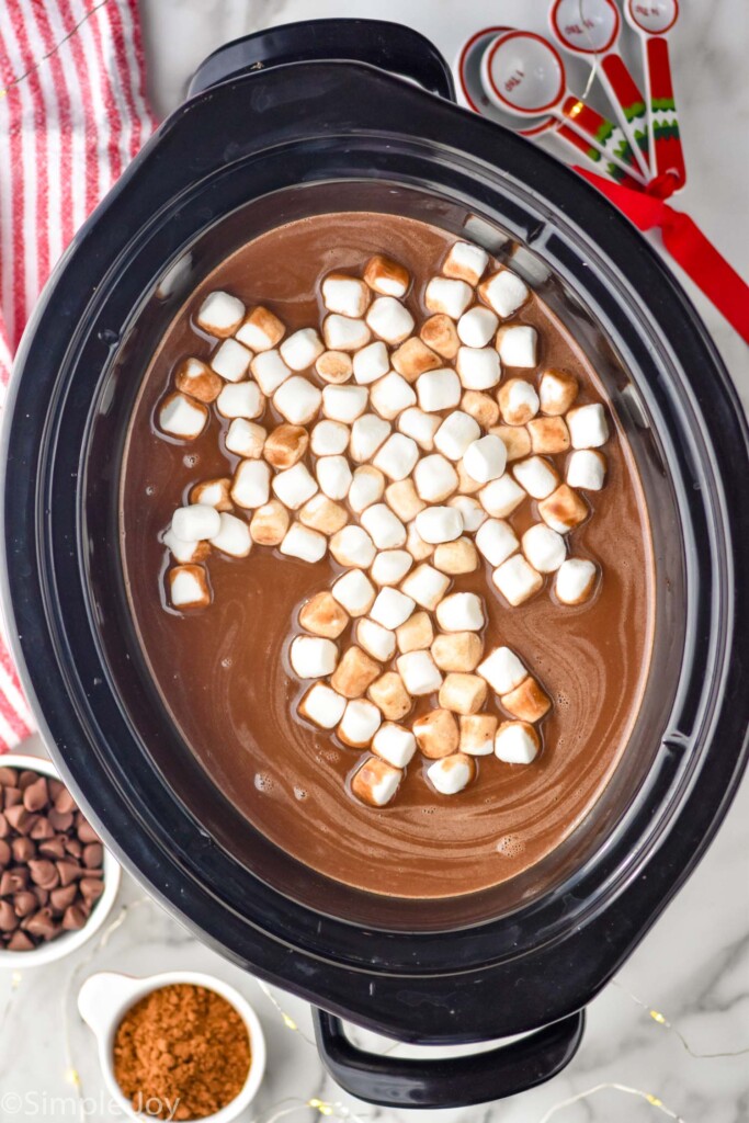 Overhead photo of crockpot hot chocolate in a 6 quart crockpot with mini marshmallows in it and cocoa powder and chocolate chips in the background
