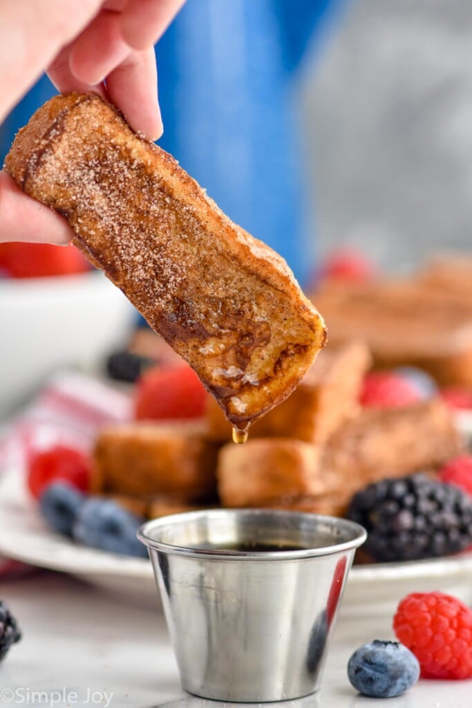 Man's hand dipping french toast stick in maple syrup. Platter of french toast sticks and fresh berries sitting in background