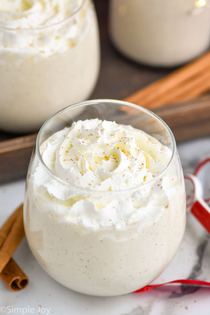 Overhead photo of homemade eggnog with whipped cream in a stemless wine glass with sparkling lights in background