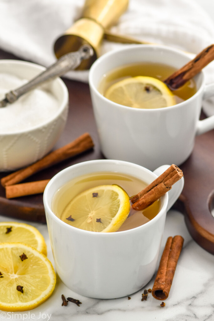 Overhead photo of two Hot Toddies in white mugs with a cinnamon stick and lemon slice in the mug and around the mug and a bowl of sugar