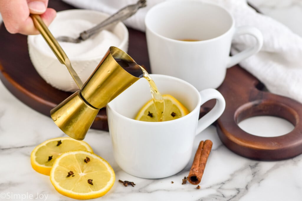 How to make a hot toddy image of liquor being poured into a white mug that has lemon in it with a lemon slice and another white mug in the background