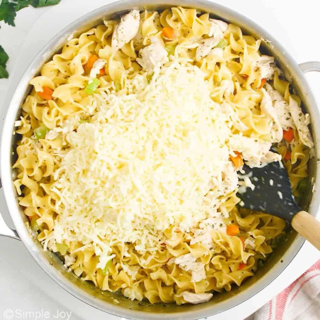 shredded cheese being mixed into noodles and vegetables in a skillet