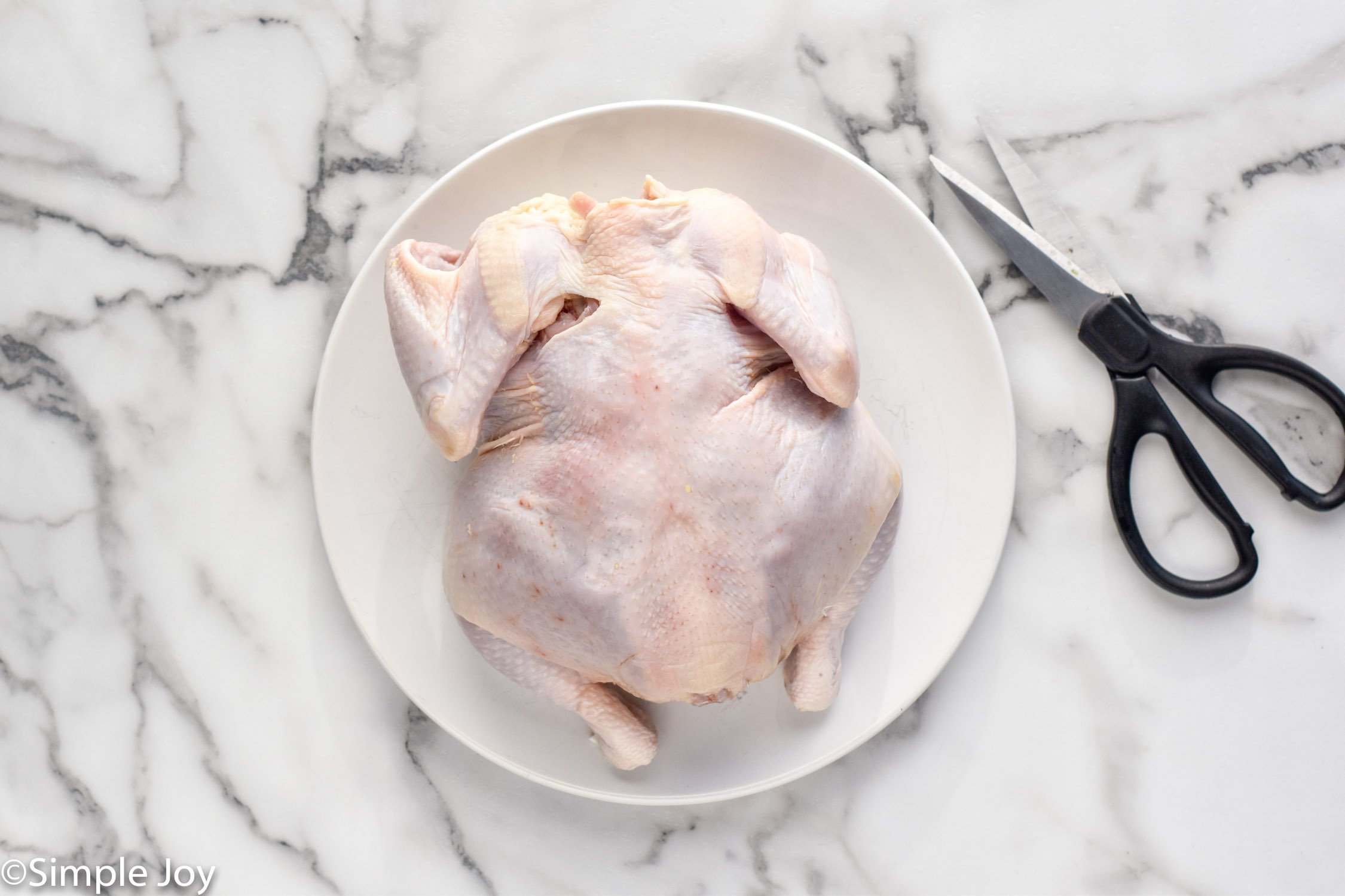 Overhead of raw chicken sitting on plate with kitchen shears beside to make spatchcock chicken recipe