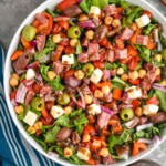 Overhead photo of Italian Chopped Salad in a white bowl with whole tomatoes, a wooden serving spoon, and dressing in the background