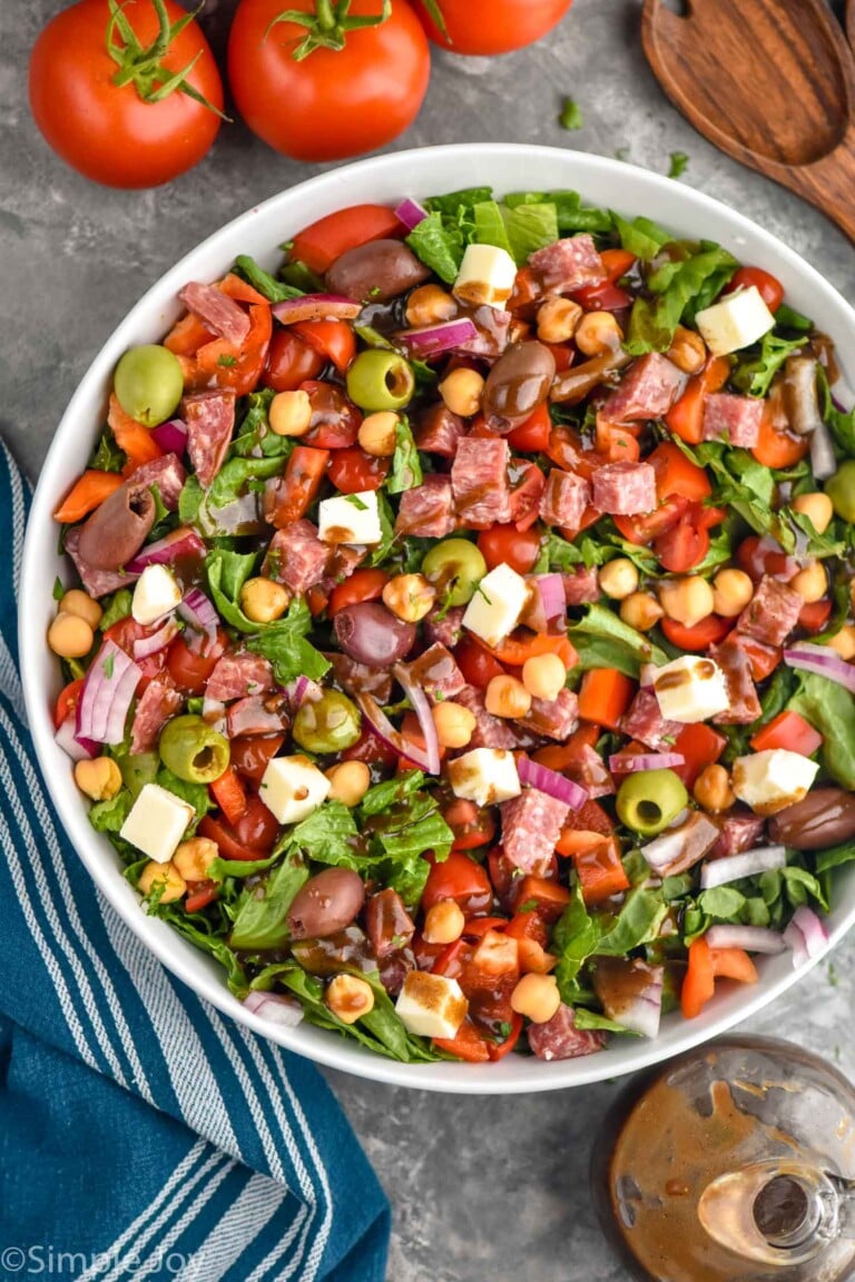 Overhead photo of Italian Chopped Salad in a white bowl with whole tomatoes, a wooden serving spoon, and dressing in the background