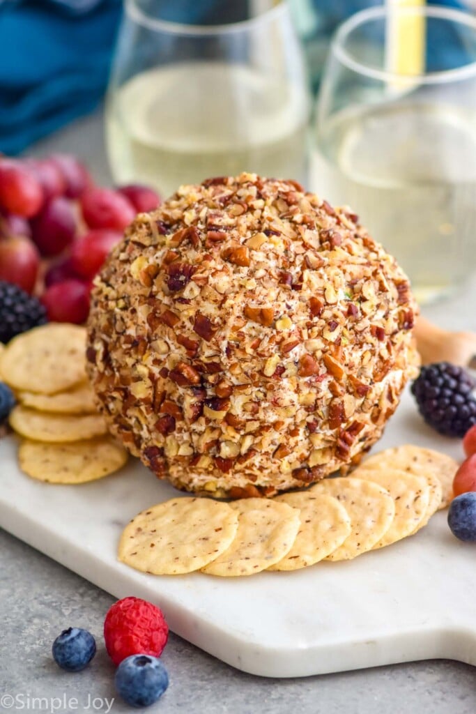 pineapple cheese ball on a serving board surrounded by crackers and fresh berries. Two glasses of white wine sit in background